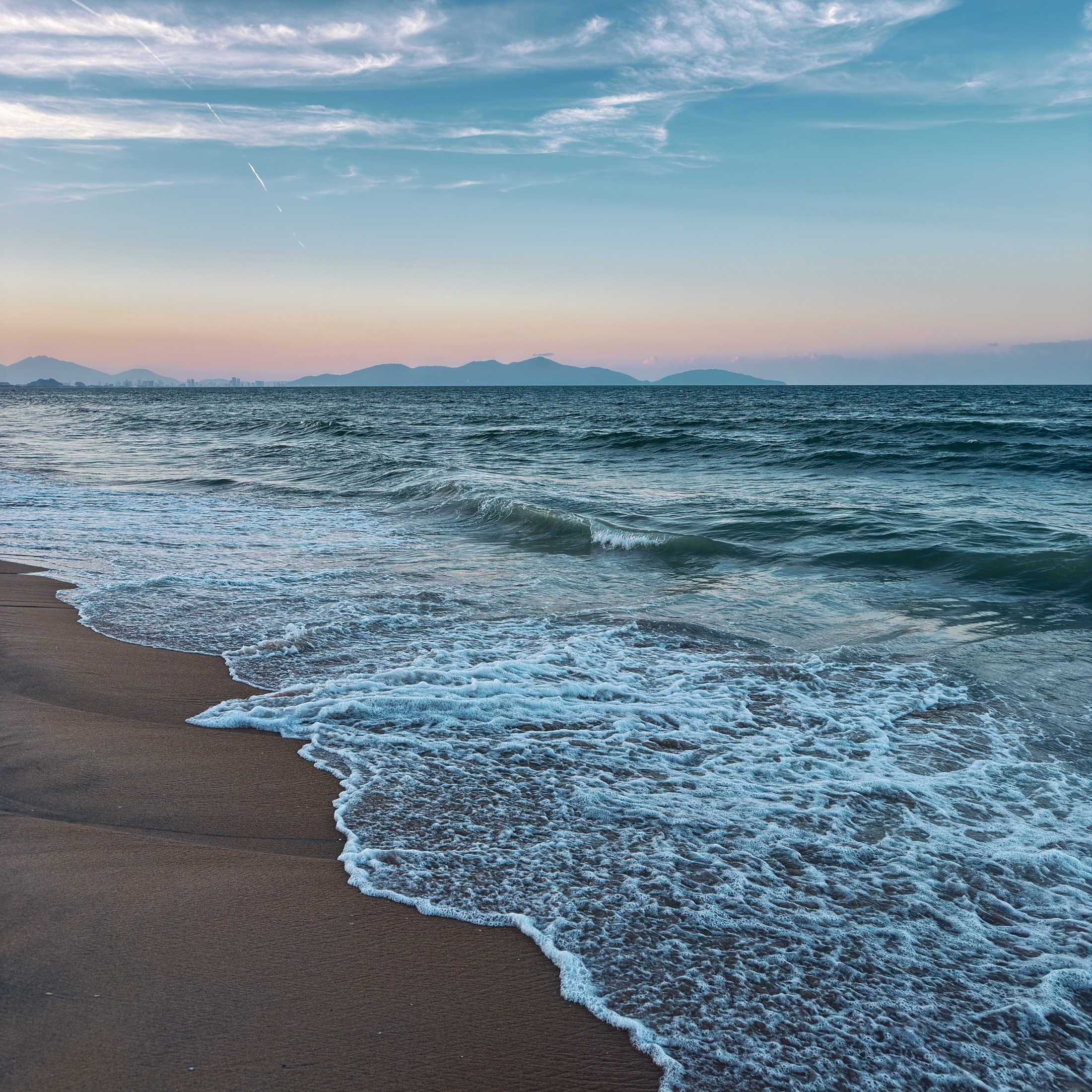 Waves breaking on a shoreline at dusk, mountains on the horizon